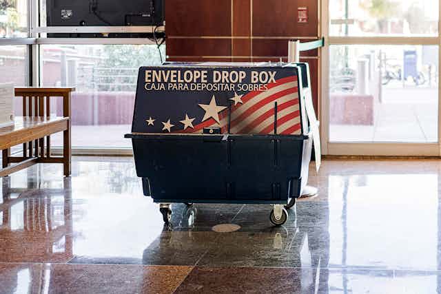 A large blue box on wheels has an American flag and says 'Envelope drop box' in what appears to be an indoor space.