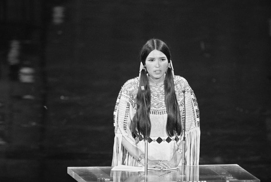 A women with long hair and in traditional Native American dress stands at a podium.