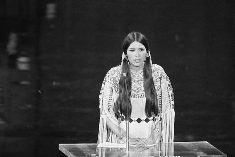 A women with long hair and in traditional Native American dress stands at a podium.