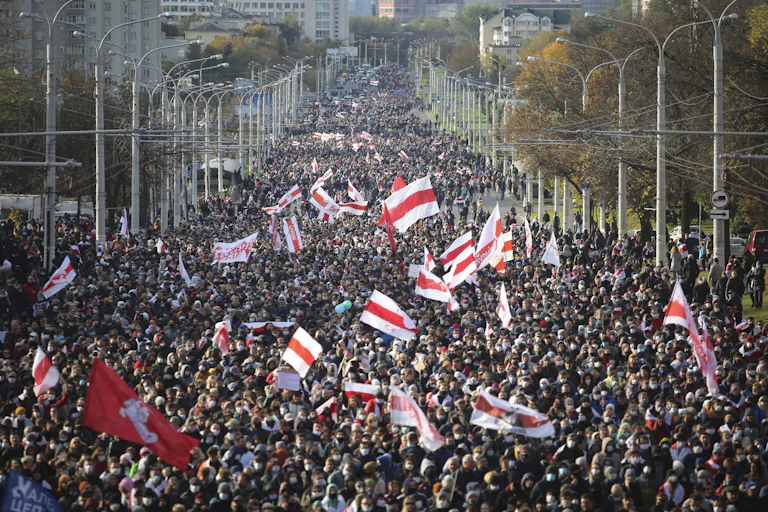 A huge crowd of people line a wide city street. Lots of red and white flags are seen.