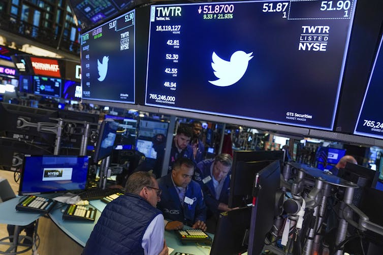 the Twitter bird logo appears in white on a large dark screen as people making trades at a stock exchange wander underneath