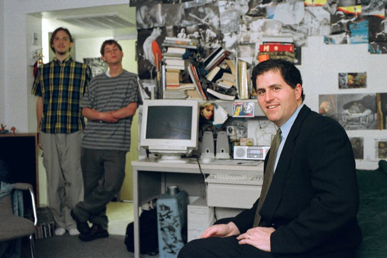 A white man in a suit sits in front of an old computer in a dorm room, while two others in business attire stand in the doorway
