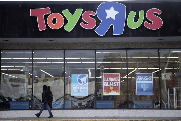 a person in a coat walks past the front of a store with a Toys r Us sign over glass doors and signs reading clearance sale