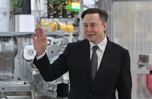 A white man in a suit holds up his right hand in front of car machinery