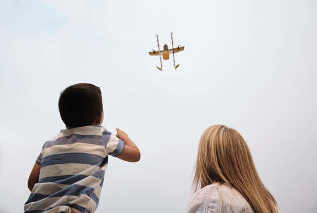 A child and blonde woman watching a drone approach in the sky