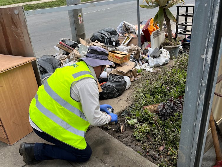 A man in a hi-viz vest kneels down to collect a sample from a nature strip