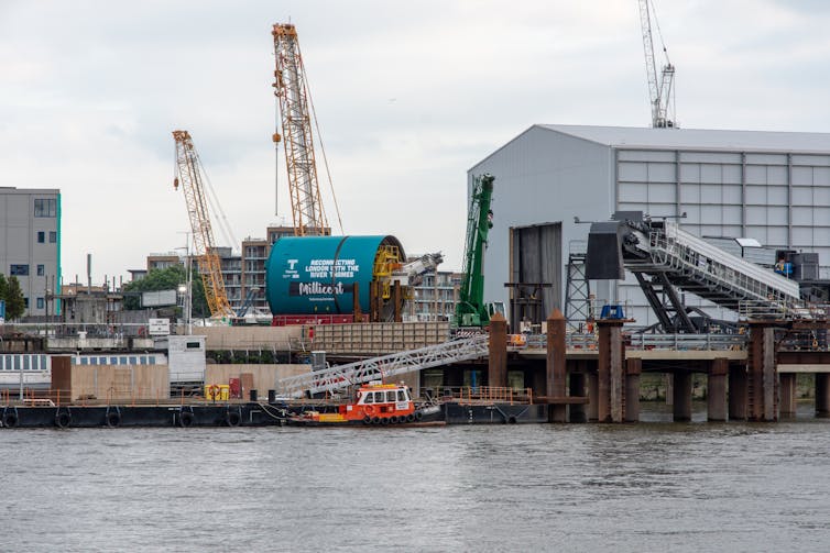 A tunnel boring machine standing on the bank of the Thames, next to a large warehouse.