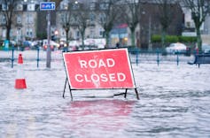 A red road closed sign emerging from a heavily flooded town road.