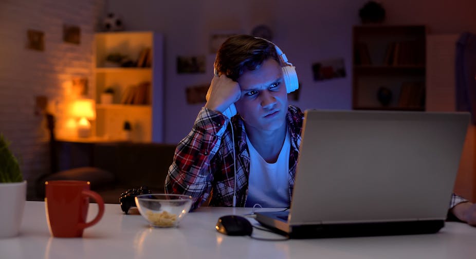 A young man wearing headphones looks angrily at a computer screen