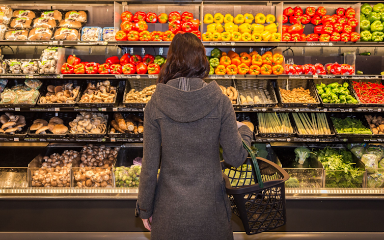 A woman stands with her back to the camera, facing a row of produce in the grocery store