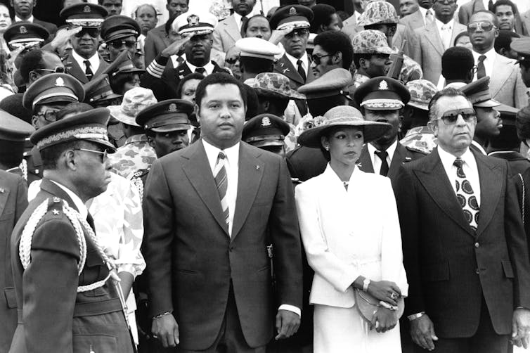 A young man in a suit stands stiffly with a military officer by his side.