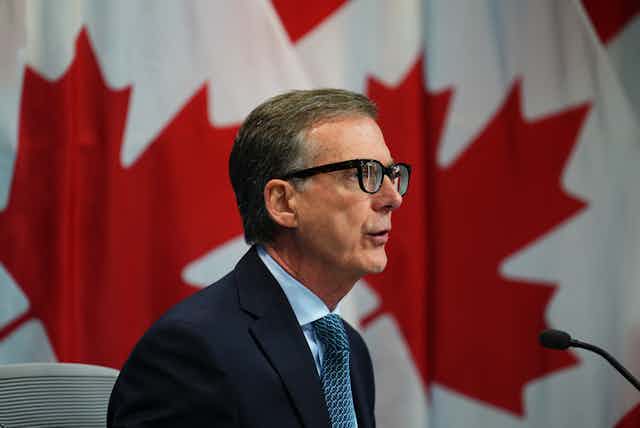 Man in glasses and a navy blazer sits in front of a Canadian flag