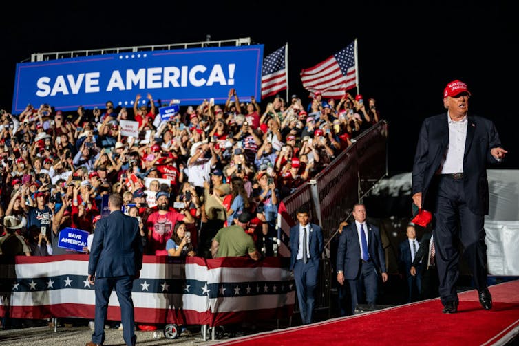 A middle-aged white man dressed in a business suit and wearing a red baseball cap walks on stage as hundreds of people in nearby grandstands applaud in support.