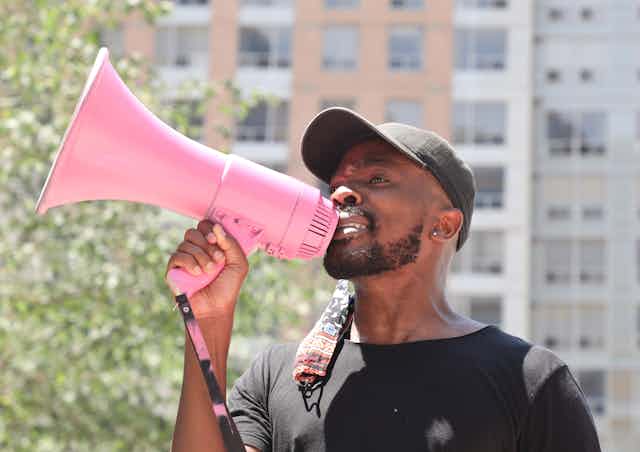 A Black man speaks into a pink magaphone.