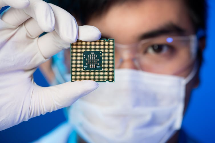 Someone wearing a mask and protective goggles holding a computer microchip in front of their face