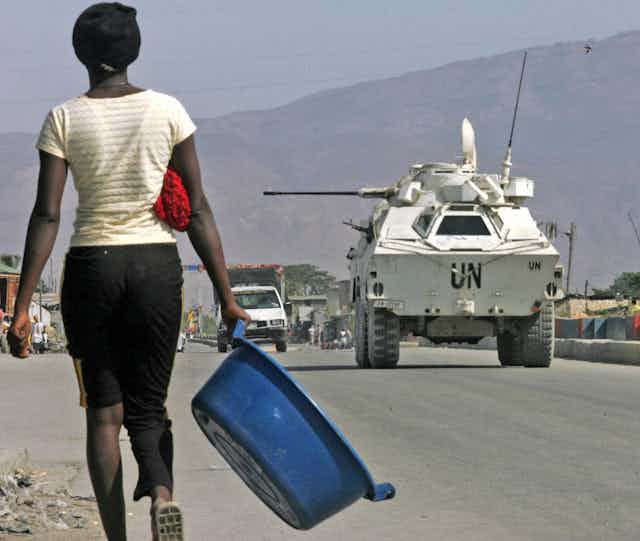 A woman carries a washbasin past a U.N. tank with mountains in the background.