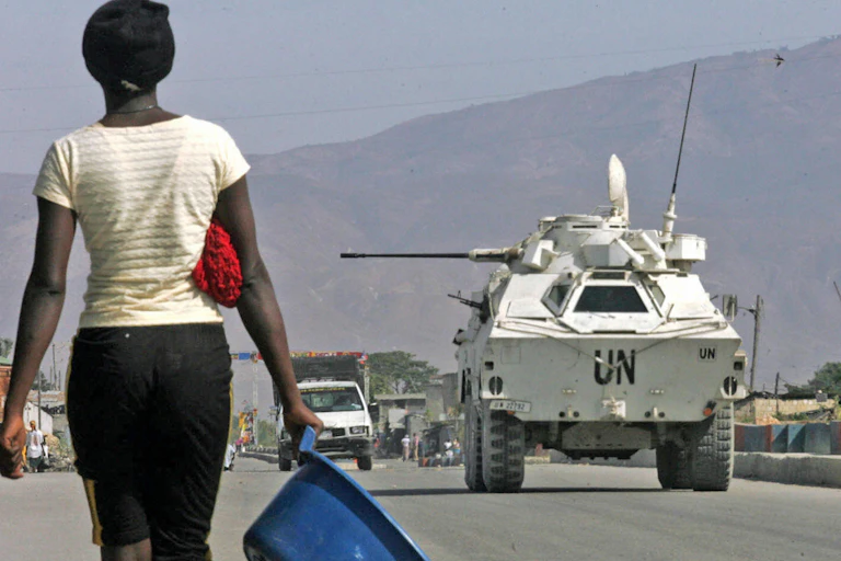 A woman carries a washbasin past a U.N. tank with mountains in the background.