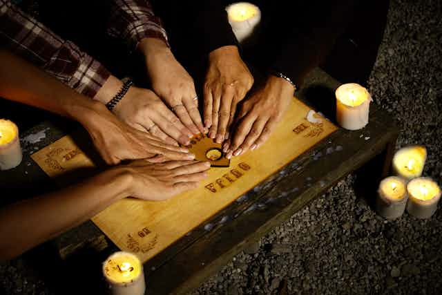 Three people have their hands placed on the planchette in order to use a Ouija board.
