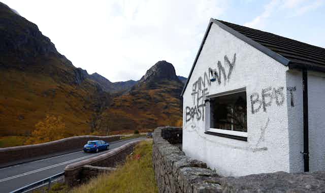 A graffitied white cottage on the side of a road with rising hills all around. 