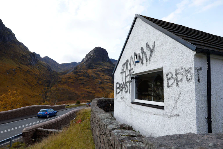 A graffitied white cottage on the side of a road with rising hills all around.