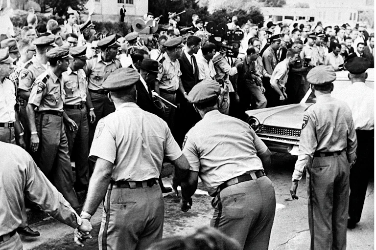 Policemen holding back a jeering crowd as a car drives through it.