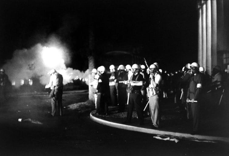 Armed troops along a sidewalk in the night, with fire in the background.