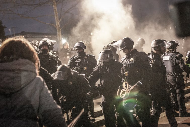Policías con cascos y equipo antidisturbios con humo de fondo.