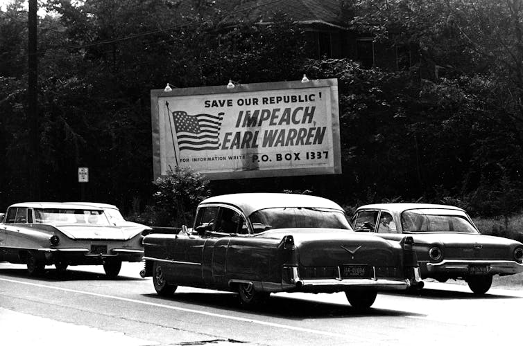 Jalan raya dengan mobil tua di atasnya dan papan iklan bertuliskan 'IMPEACH EARL WARREN' di sampingnya.