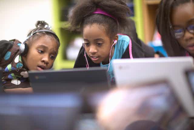 Three young Black girls look at one of two computer screens. Two are wearing headphones.