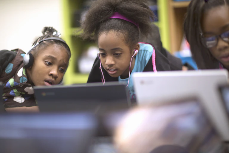 Three young Black girls look at one of two computer screens. Two are wearing headphones.