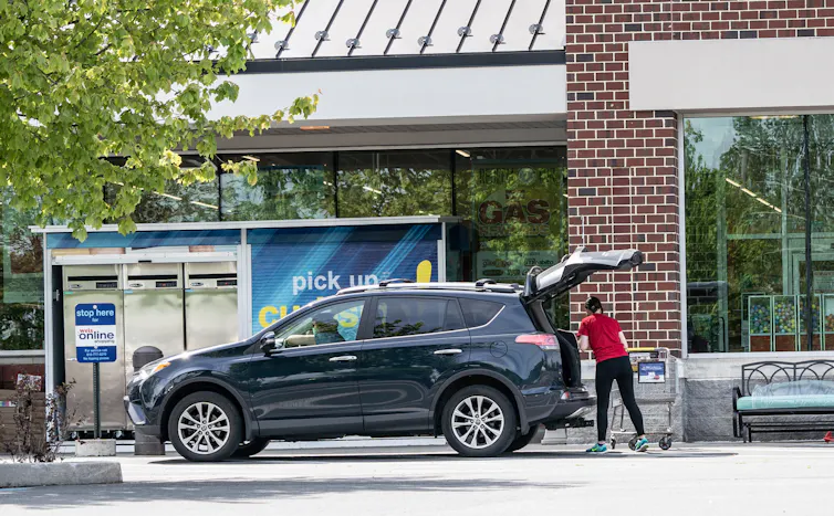 A person wearing a red T shirt puts groceries into the back of a black car.