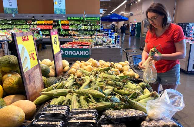 woman in red shirt wearing glasses puts corn cobs in a plastic bag in the produce aisle