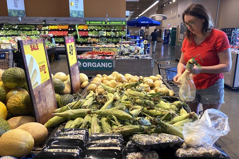 woman in red shirt wearing glasses puts corn cobs in a plastic bag in the produce aisle