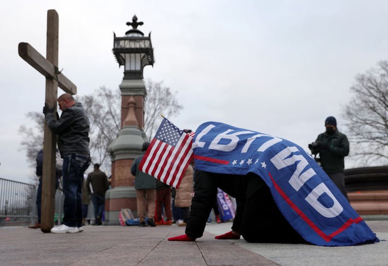 Someone draped in a flag that says 'Trump,' holding an American flag, kneels before a man holding a large cross outside on a sidewalk.