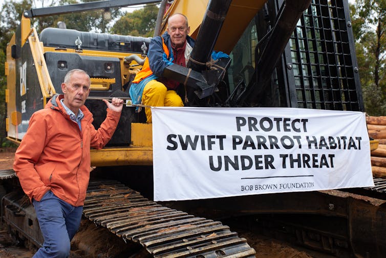 two men with a bulldozer and sign reading 'Protect swift parrot habitat under threat'