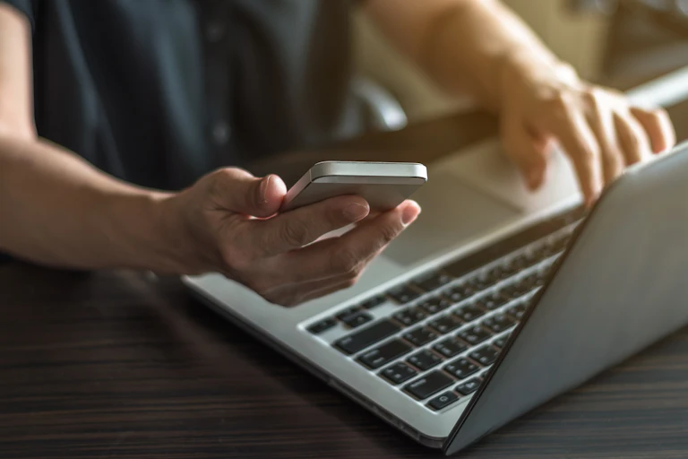 Someone at a laptop holds up a phone.