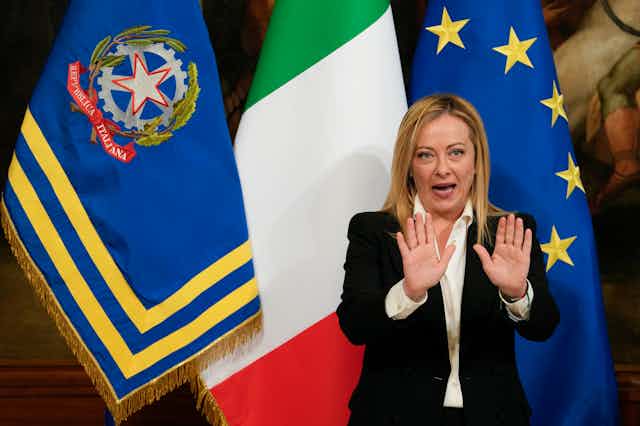 A blonde woman stands in front of three different flags, including the green, white and red Italian flag, with her hands held up in front of her as she speaks.