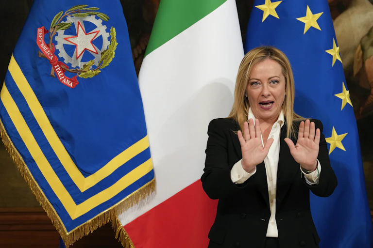 A blonde woman stands in front of three different flags, including the green, white and red Italian flag, with her hands held up in front of her as she speaks.
