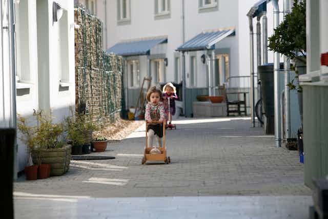 Two little girls scoot down a pathway between white and grey houses. 