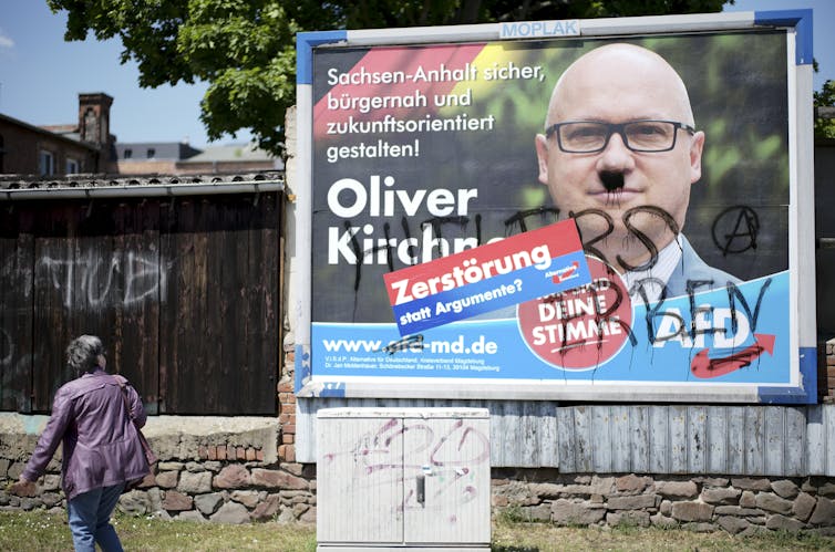 A vandalized campaign poster shows a candidate with a Hitler moustache.