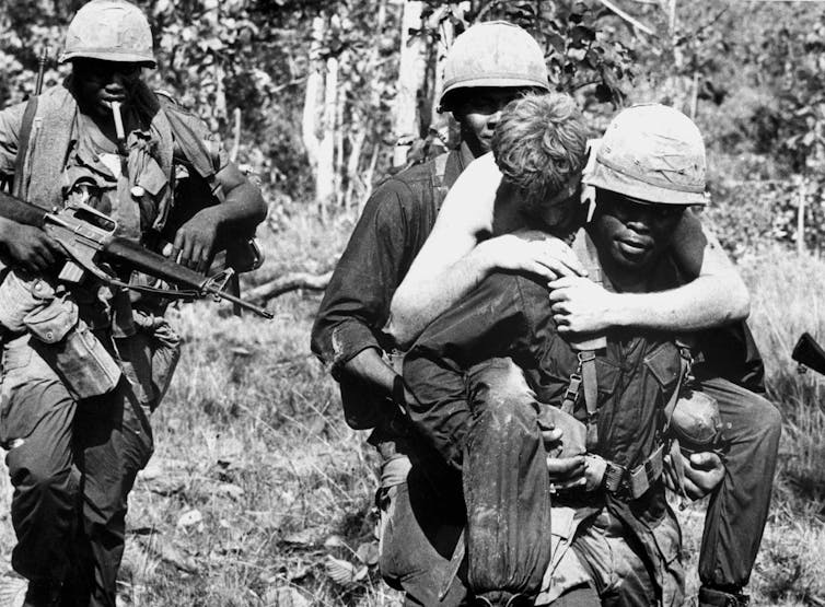 A wounded white soldier is carried by a Black soldier during the Vietnam War.