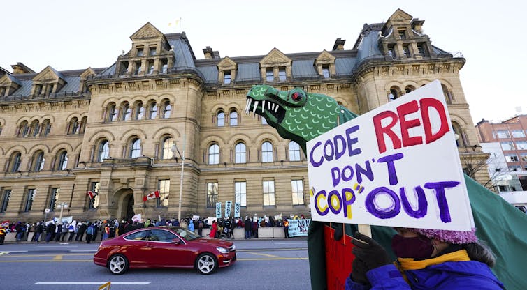 A person carrying a sign that says code red, don't cop out stands in front of a stone building with other protesters