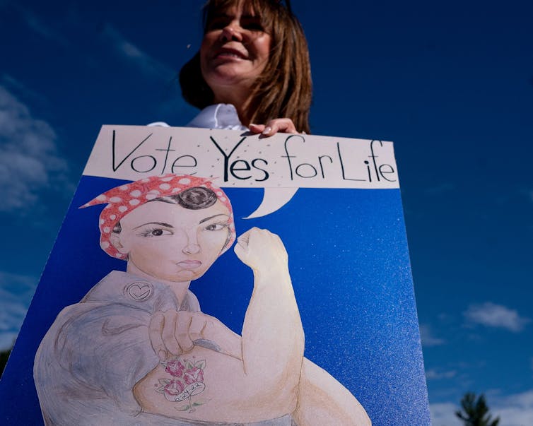 A white woman with brown hair holds up a Rosie the Riveter poster that says 'Vote yes for life'
