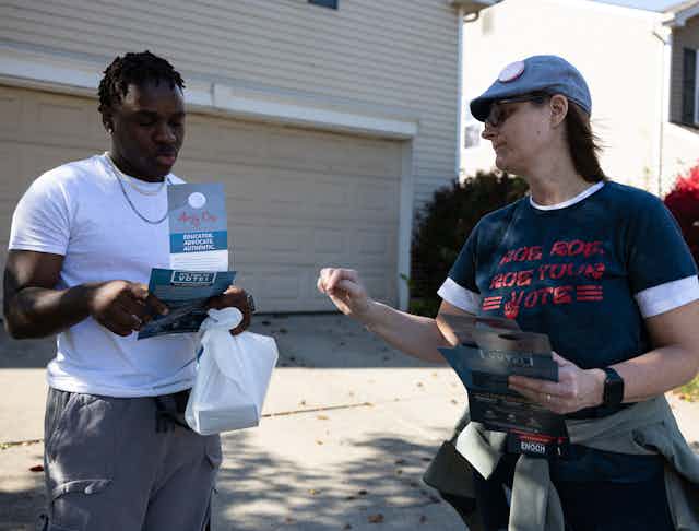 A woman wearing a hat and a blue shirt that says 'Roe roe roe your vote, speaks with a black young man holding campaign information outside of a beige house, standing in a driveway.