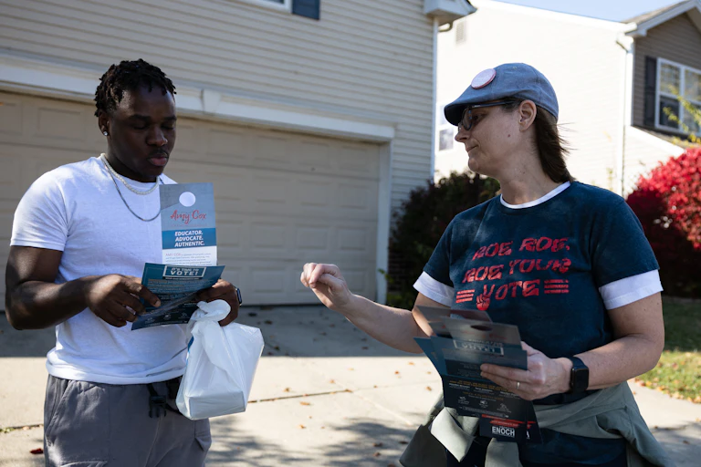 A woman wearing a hat and a blue shirt that says 'Roe roe roe your vote, speaks with a black young man holding campaign information outside of a beige house, standing in a driveway.