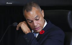 A man in a dark suit with a red poppy on his lapel wipes a tear from his face with the back of his hand.