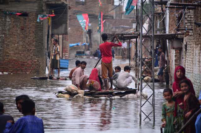 Groups of men on makeshift rafts float down a flooded street while people stand in flooded doorways.