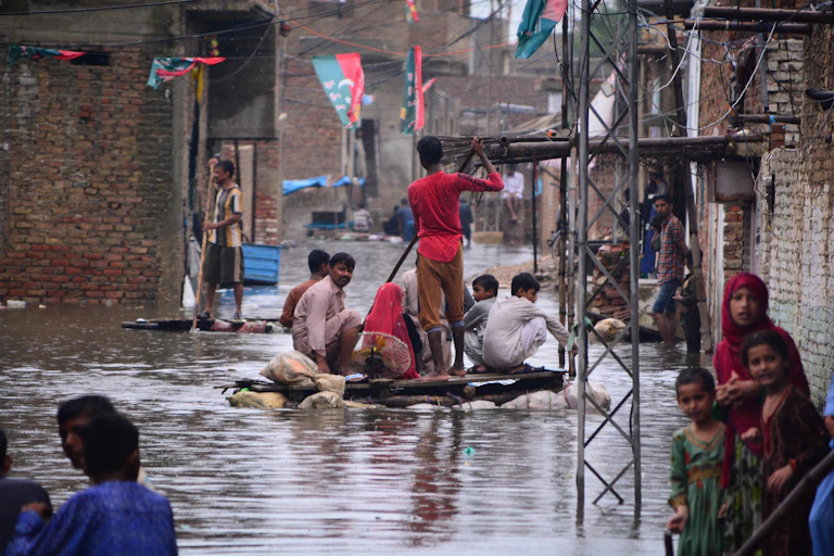 Groups of men on makeshift rafts float down a flooded street while people stand in flooded doorways.