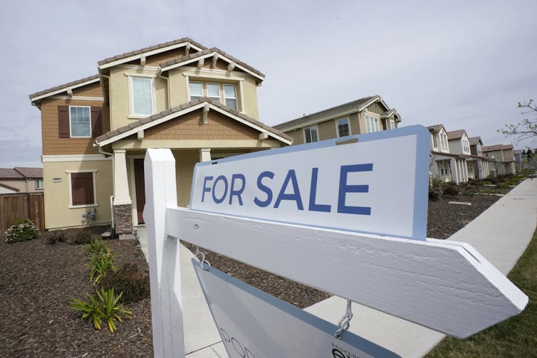 For sale sign sits atop a white post in front of a cream/brown multi-floor house