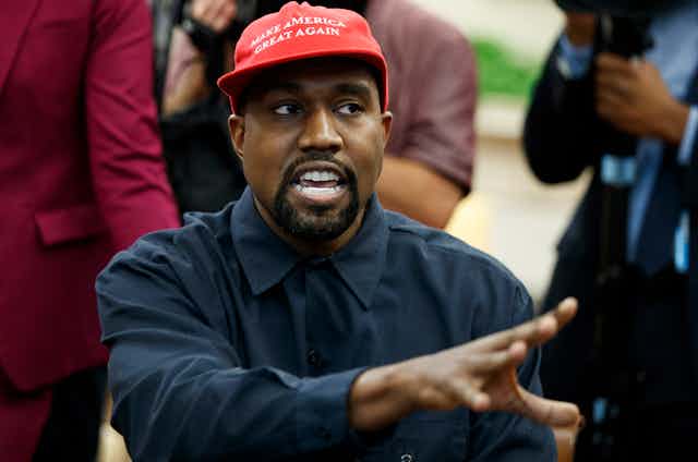 a black man wearing a red make America great hat and a blue dress shirt gestures with his right hand